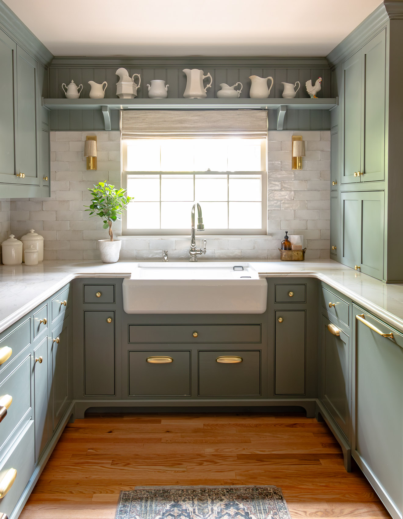 Green kitchen with white farmhouse sink beneath a window, brass hardware, and blue-green cabinets with a shelf of white teapots above the window.