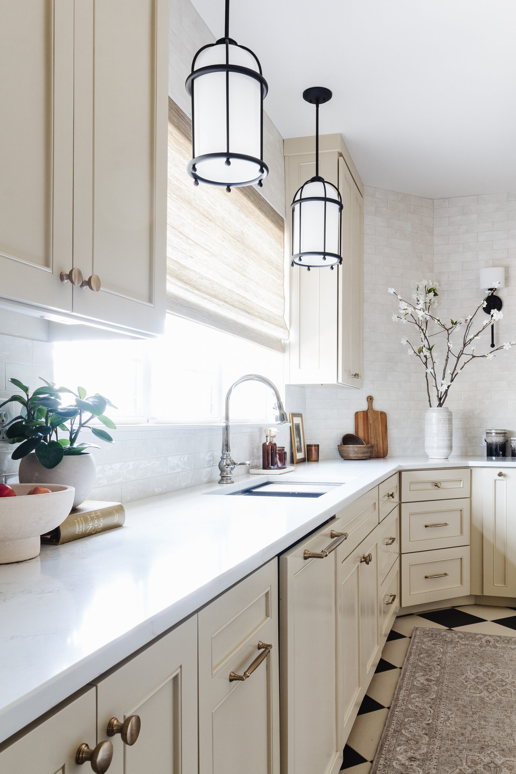 Cream kitchen with marble countertop, beige cabinetry, and two black metal pendant lights over the counter. A potted plant sits by the window and a checkered floor is visible.