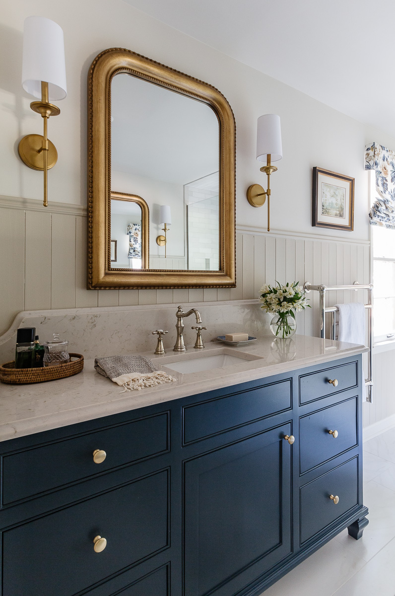 Navy bathroom vanity with a marble countertop, brass hardware, and a large ornate gold-framed mirror flanked by brass wall sconces.