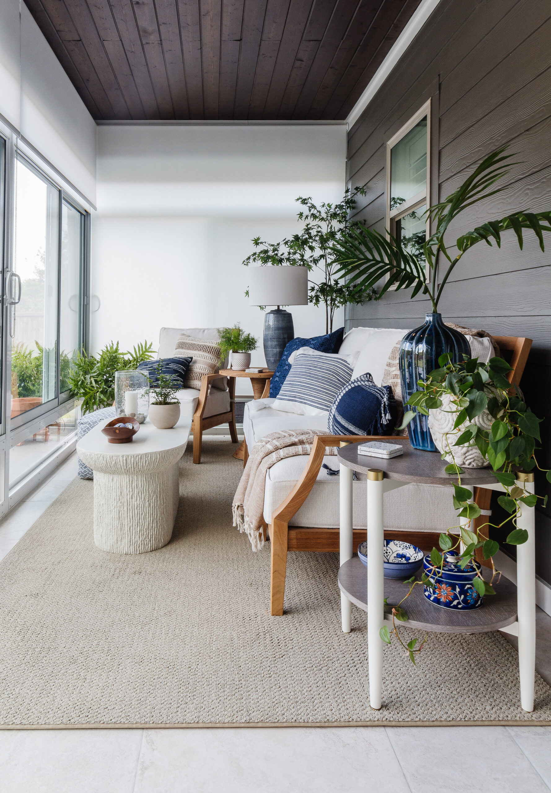 Bright sunlit living room with beige sofa, blue cushions, wooden furniture, and many potted plants by a large window.