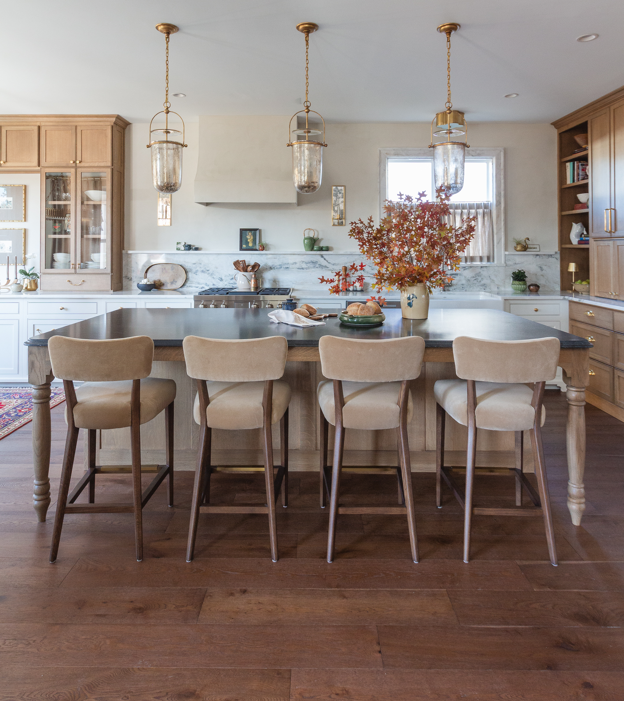 Kitchen with marble island, six beige stools, warm wood cabinets, and three brass pendant lights overhead.